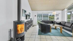 Een moderne woonkamer met een houtkachel, zwarte leren banken, een houten salontafel en een groot raam met uitzicht op de natuur.