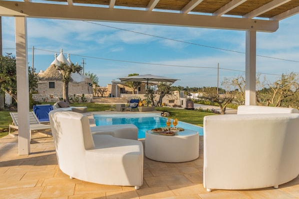 Zona de piscina con sillas de lounge blancas, una mesa redonda con comida y bebidas, y un edificio de piedra al fondo.
