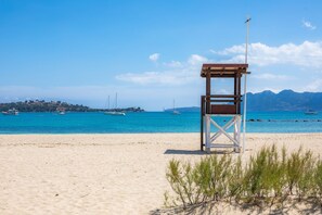 Ein Strand mit einem Rettungsturm, klarem blauen Wasser, Booten und Bergen im Hintergrund.
