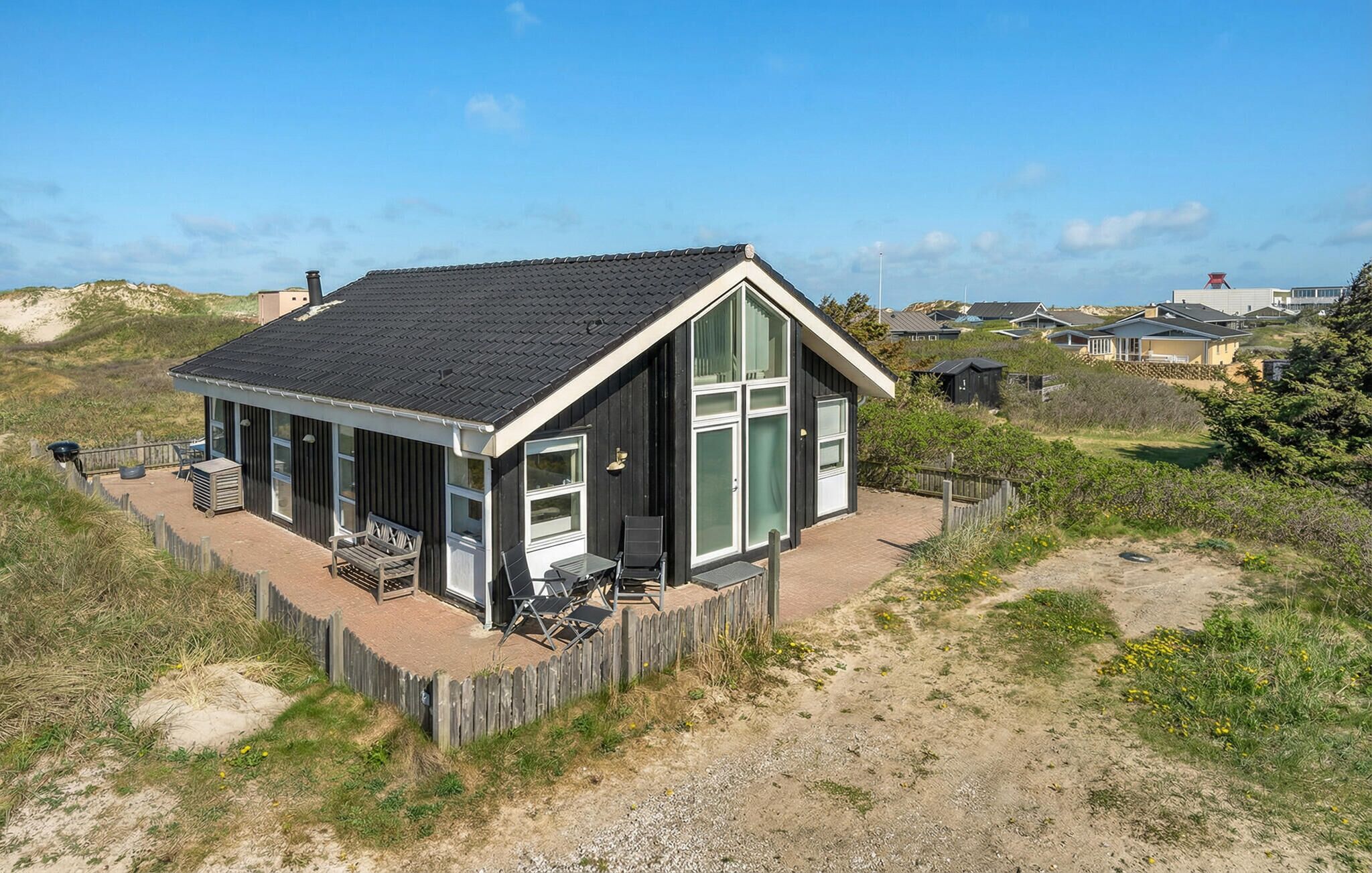 A coastal house with a deck, large windows, and a sandy surrounding.