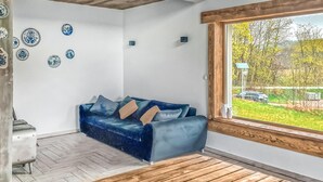 Un salon moderne avec un canapé bleu, un plancher en bois et une grande fenêtre donnant sur un paysage verdoyant et une route.