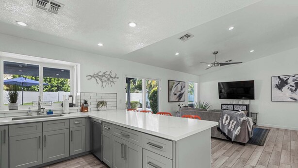 A modern kitchen with grey cabinets, a white countertop, and a large island. There is a dining area with orange chairs and a grey sofa. A ceiling fan and a flat-screen TV are visible. - Spring Song: Location, Style, Pool, Firepit, BBQ! (Scottsdale)
