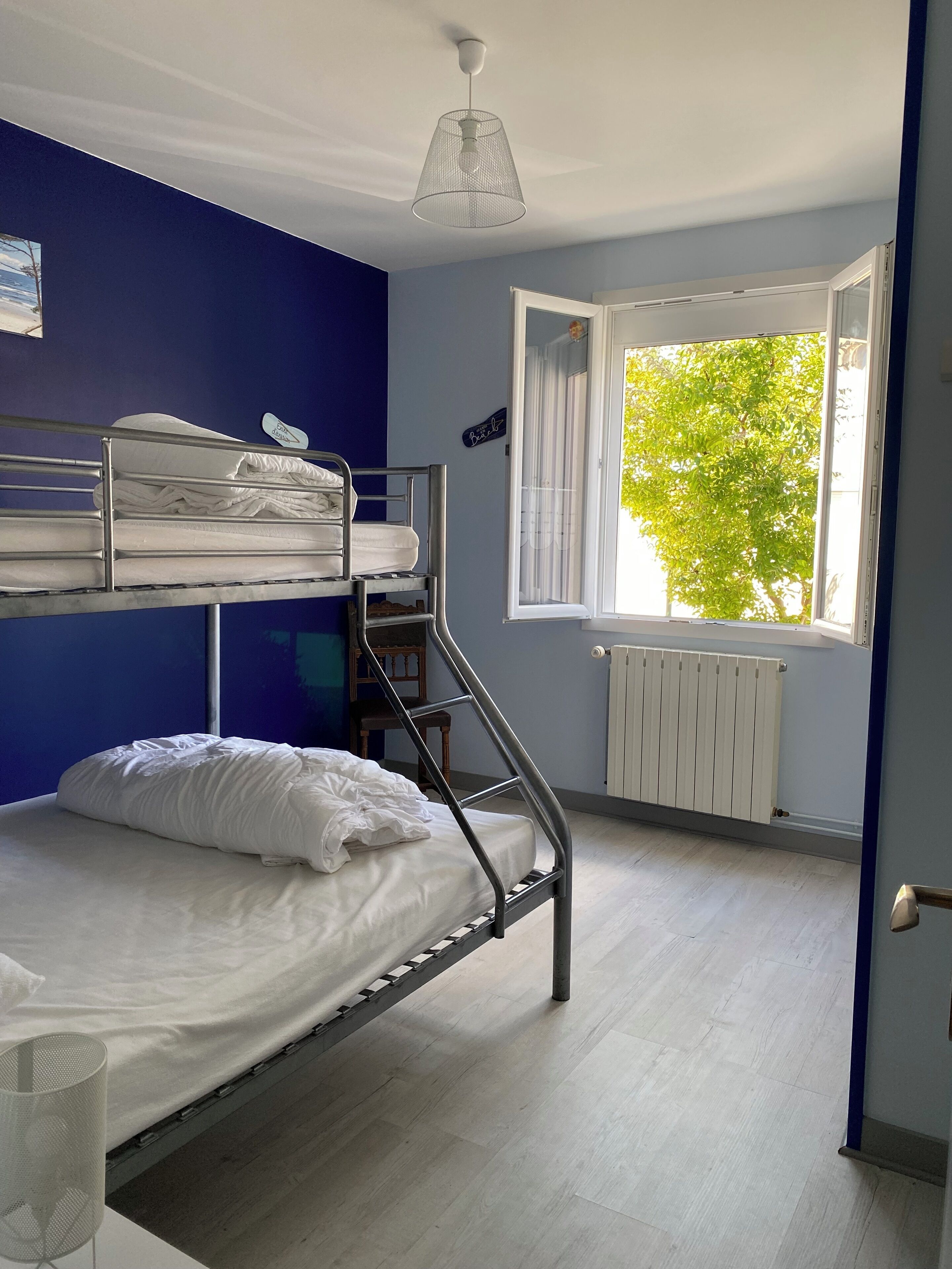 A dormitory room with bunk beds, a radiator, a window with a view of trees, and a hanging light fixture.