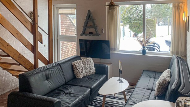A cozy living room with a black leather sofa, a white coffee table, and a TV mounted on the wall. A wooden staircase is visible to the left, and a window with a view of the outside is on the right. - 2 bedroom lovely home in Rømø (Rømø)