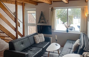 Un salon chaleureux avec un canapé en cuir noir, une table basse blanche et un téléviseur fixé au mur. Un escalier en bois est visible sur la gauche, et une fenêtre donnant sur l’extérieur se trouve sur la droite.