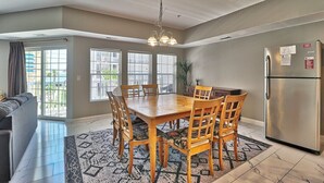 Un coin repas avec une table et des chaises en bois, un lustre, un réfrigérateur et une vue sur l’extérieur grâce à de grandes fenêtres.
