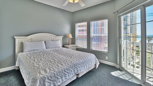 Une chambre à coucher comprenant un lit, un ventilateur de plafond, une table de chevet, deux fenêtres et une vue sur des immeubles.