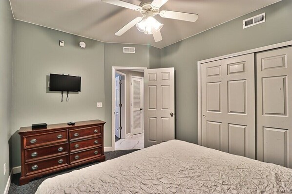 Un dormitorio con cama, cómoda, televisor, ventilador de techo y puertas blancas.