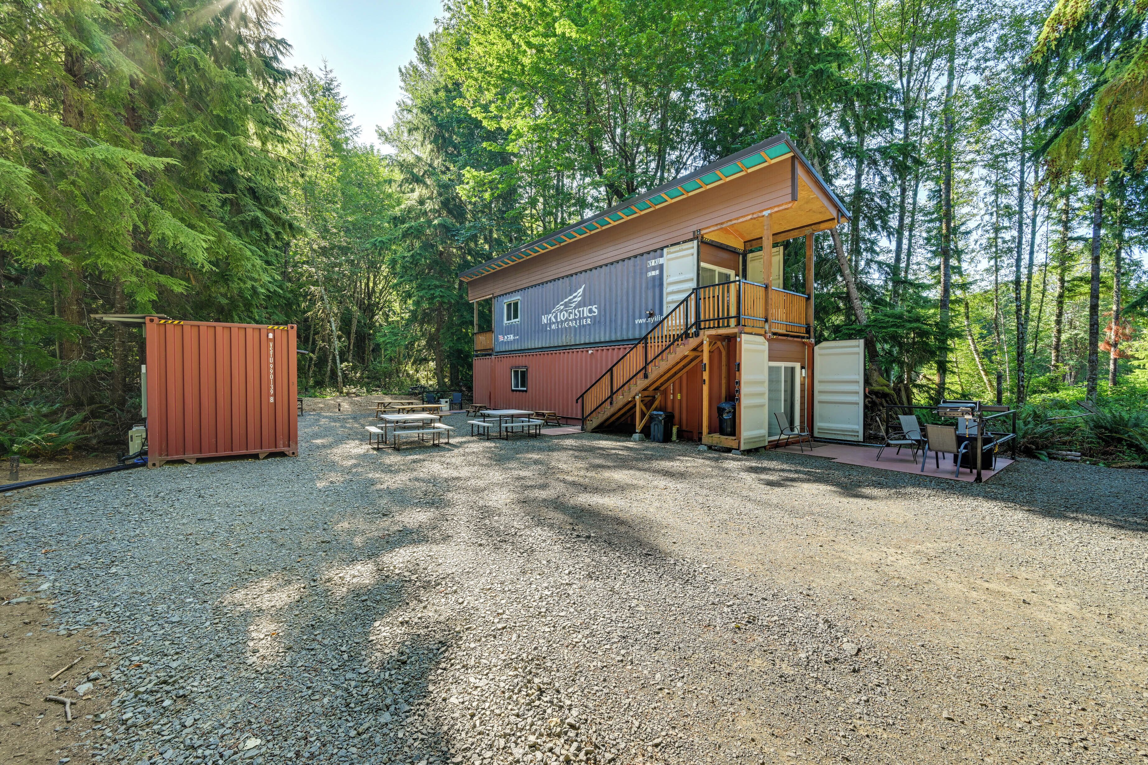 Une cabane à deux étages dotée d’une terrasse et d’un espace salon extérieur, le tout entouré de forêt.