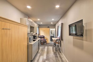 A narrow hallway with a kitchen area on the left, a seating area with a chair and table in the center, and a television mounted on the wall to the right.