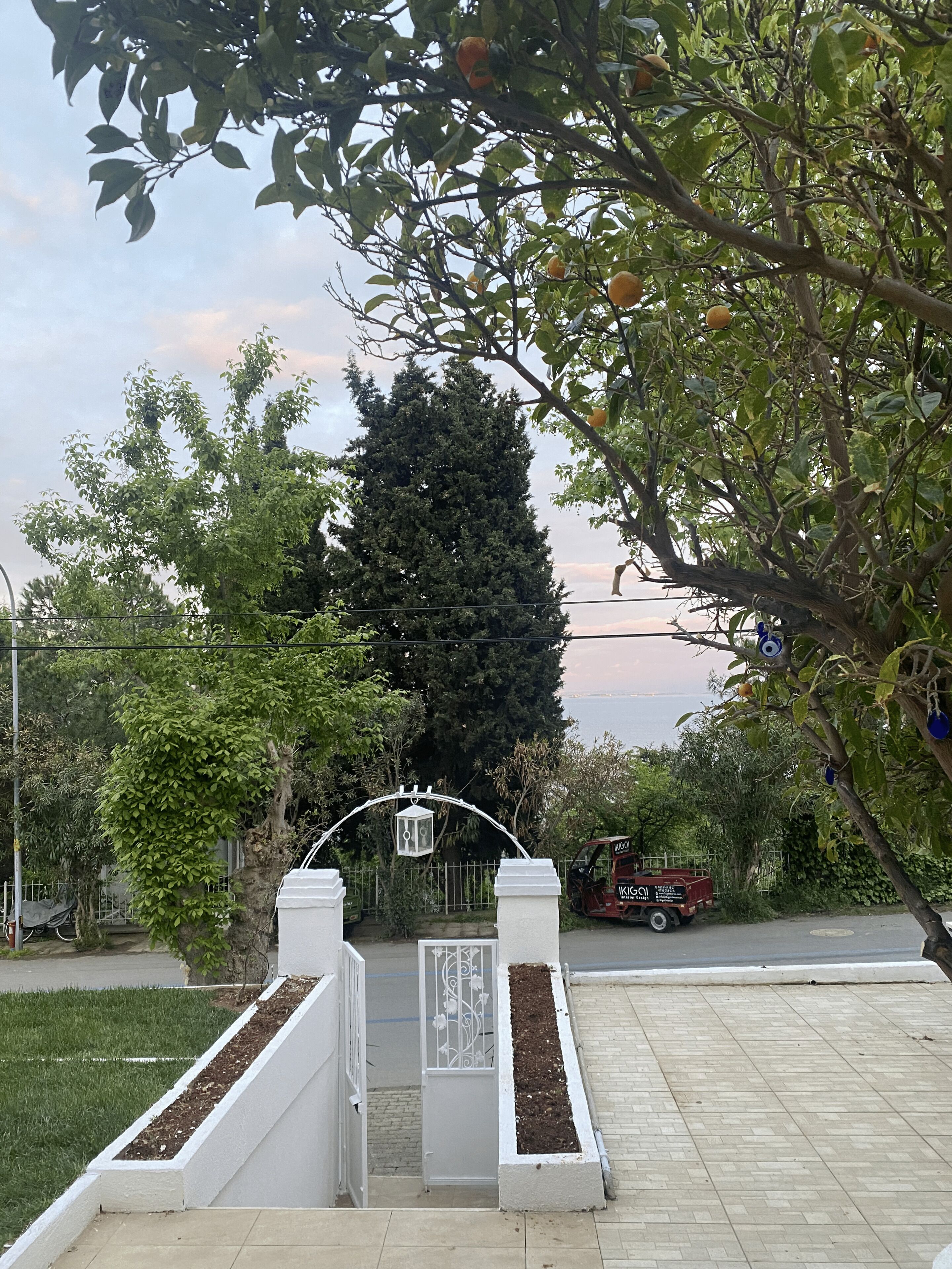 Una entrada cerrada con un arco de metal, un muro blanco con un panel decorativo y un camino pavimentado que conduce a una carretera.