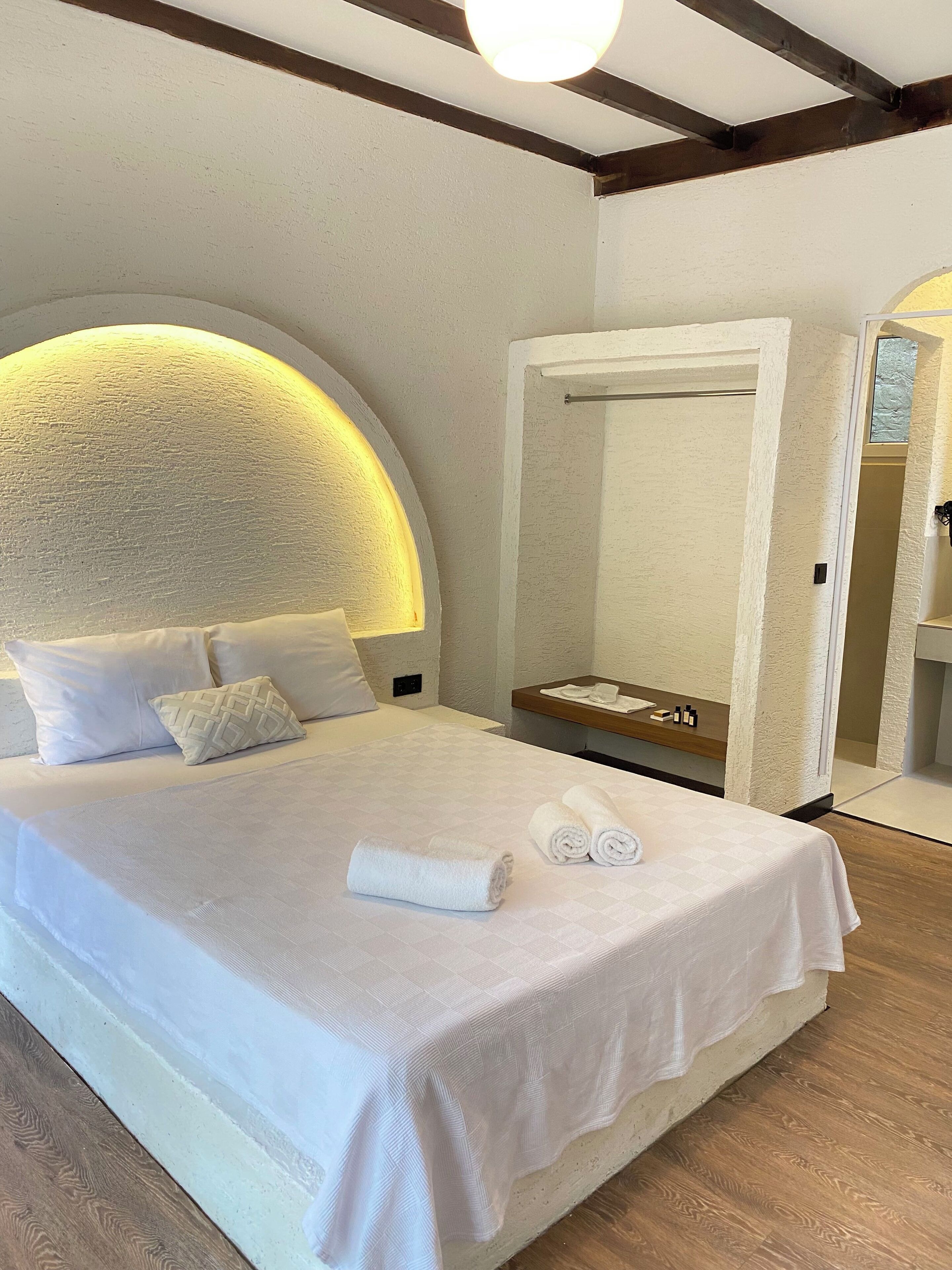 A neatly made bed with white linens and a rolled towel, a bedside table with toiletries, and a large mirror above the bed.