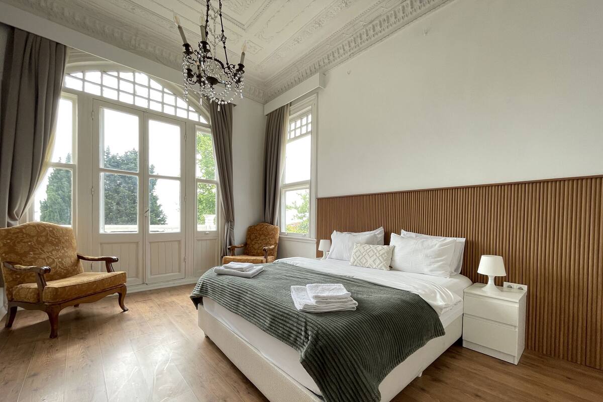 A spacious bedroom with a large bed, a wooden headboard, a chandelier, and a view of greenery through the windows.