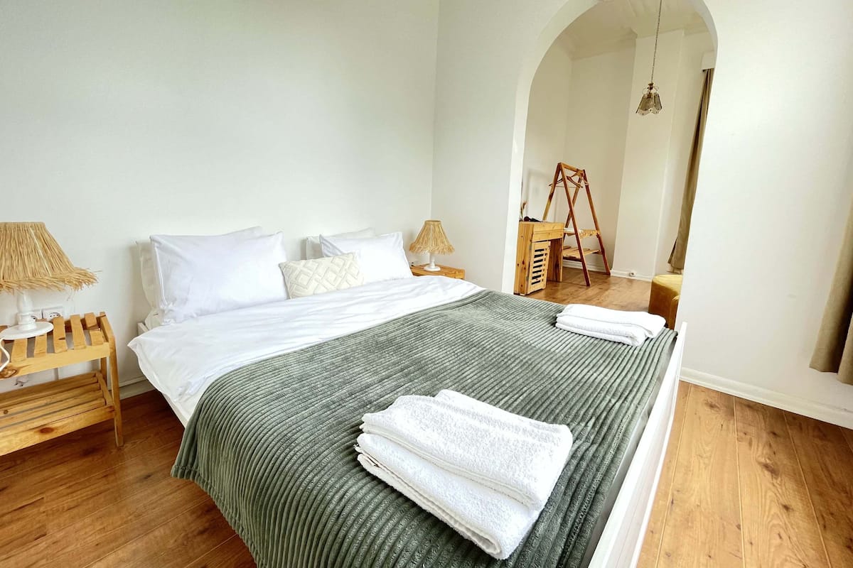 A neatly made bed with white linens and a green striped blanket, a wooden nightstand with a lamp, and a wooden ladder against the wall.