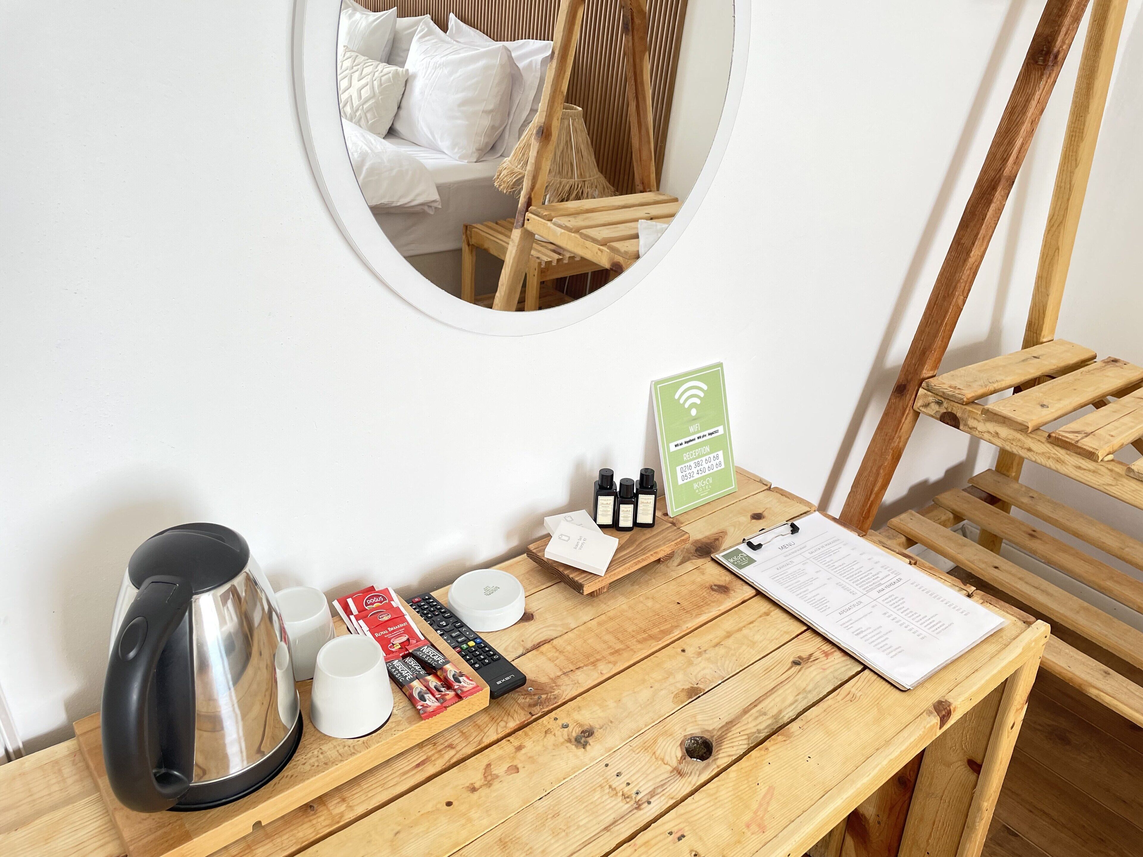Une table en bois avec une bouilloire, des tasses, une télécommande et un menu. Un miroir reflète un lit avec des oreillers.