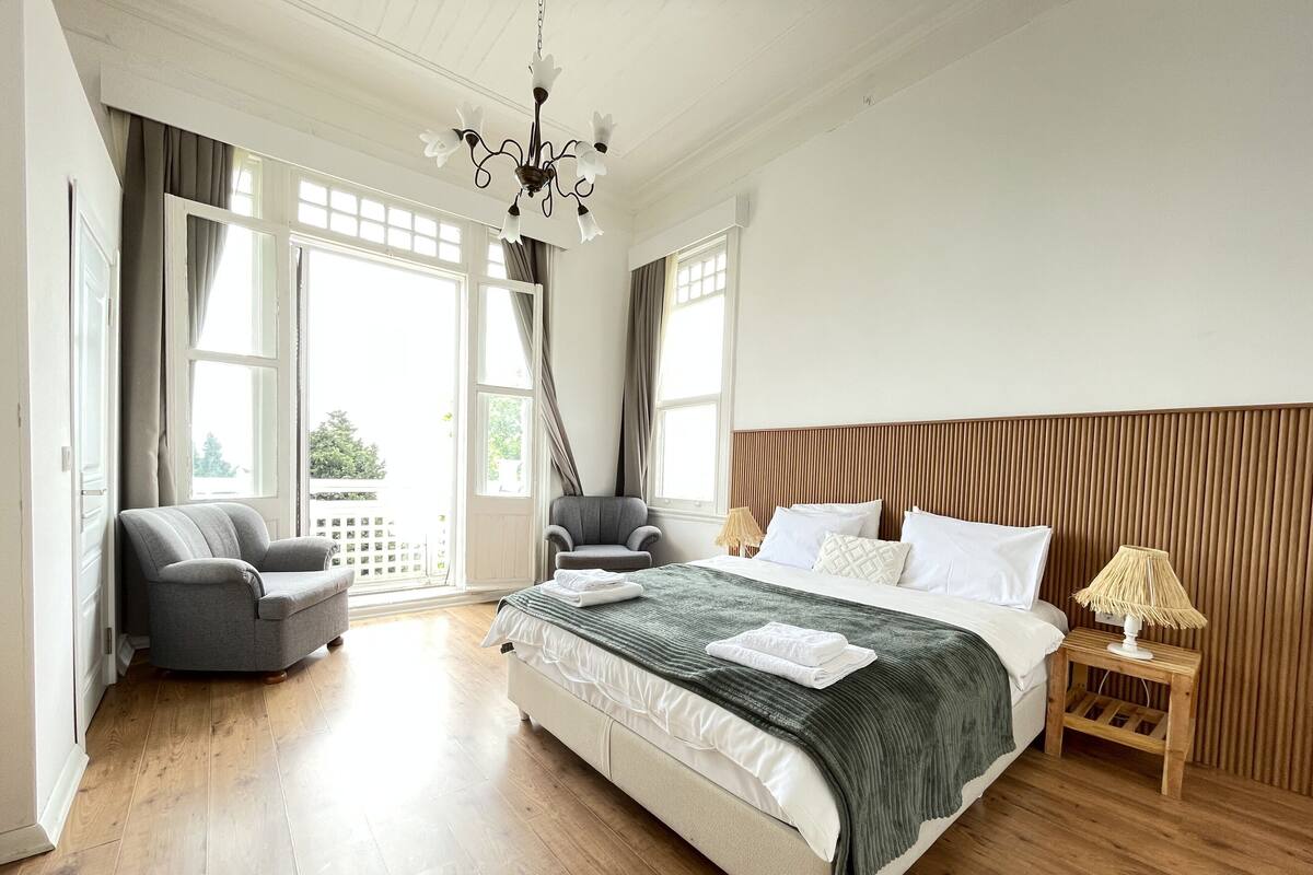 A hotel room with a large bed, a grey armchair, a wooden headboard, a nightstand with a lamp, and a view of the outdoors through large windows.