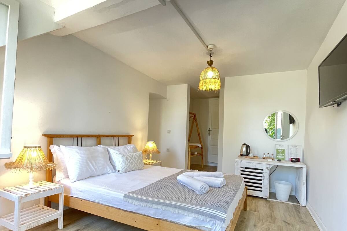 A bedroom with a wooden bed, white bedding, a bedside table with a lamp, a small desk with a mirror, and a ceiling light fixture.
