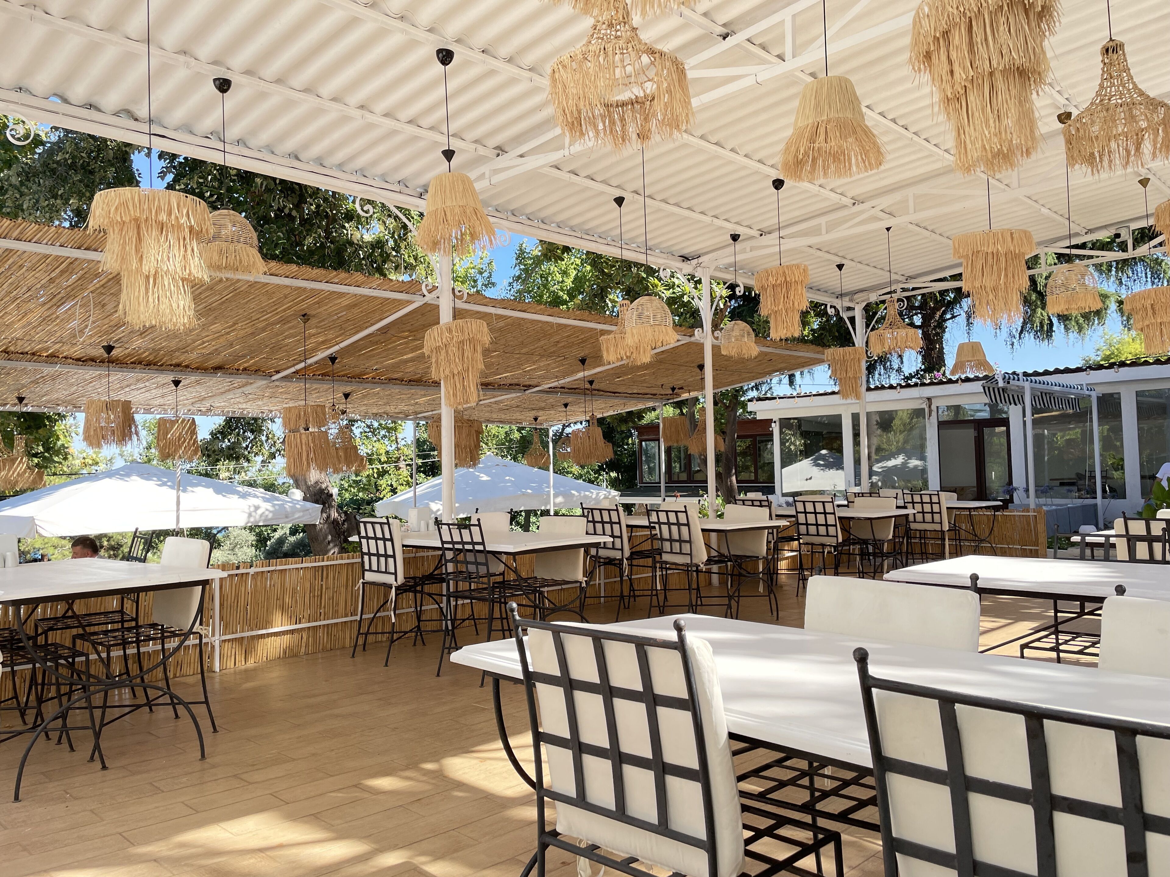 An outdoor dining area with tables and chairs, covered by a white roof with hanging rattan lamps.