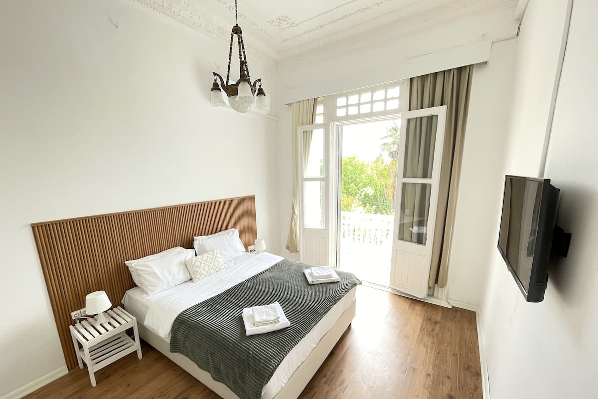 A neatly arranged bedroom with a wooden headboard, a bed with white and green bedding, a bedside table with a lamp, and a TV mounted on the wall.