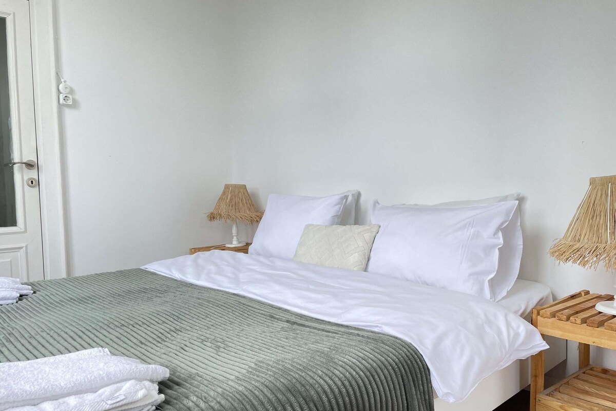 A neatly made bed with white linens and a green striped blanket, two bedside lamps, and a wooden nightstand with a woven lamp shade.
