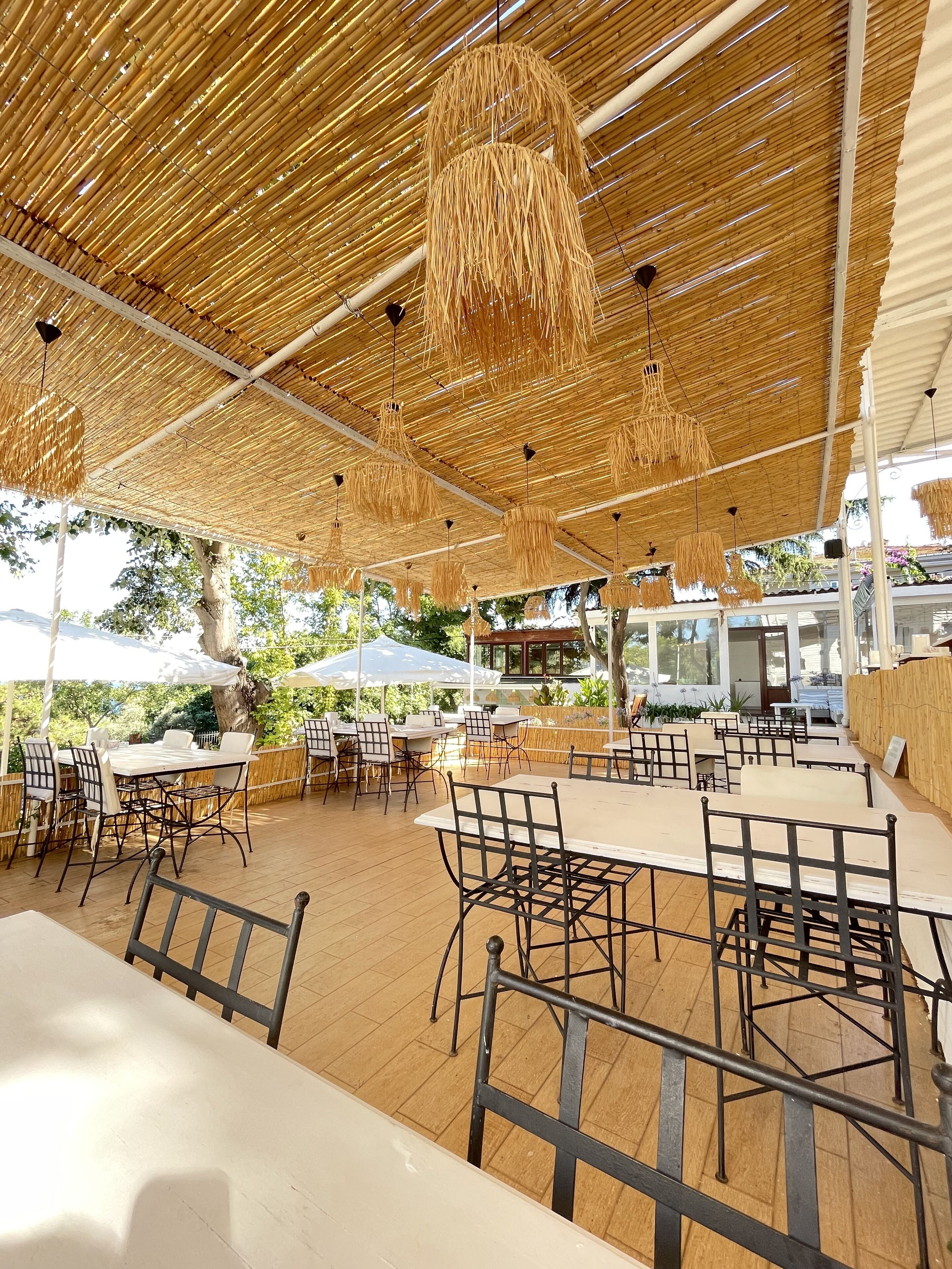 An outdoor dining area with tables and chairs, covered by a bamboo roof, and surrounded by greenery.