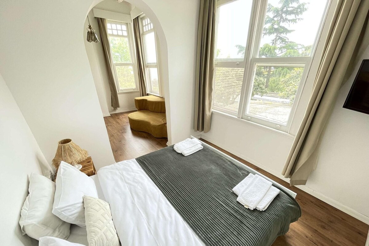 A bedroom with a bed, pillows, a bedside table, a lamp, and a window with curtains.