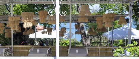 An outdoor seating area with tables and chairs, covered by a white canopy, and hanging thatched lamps.