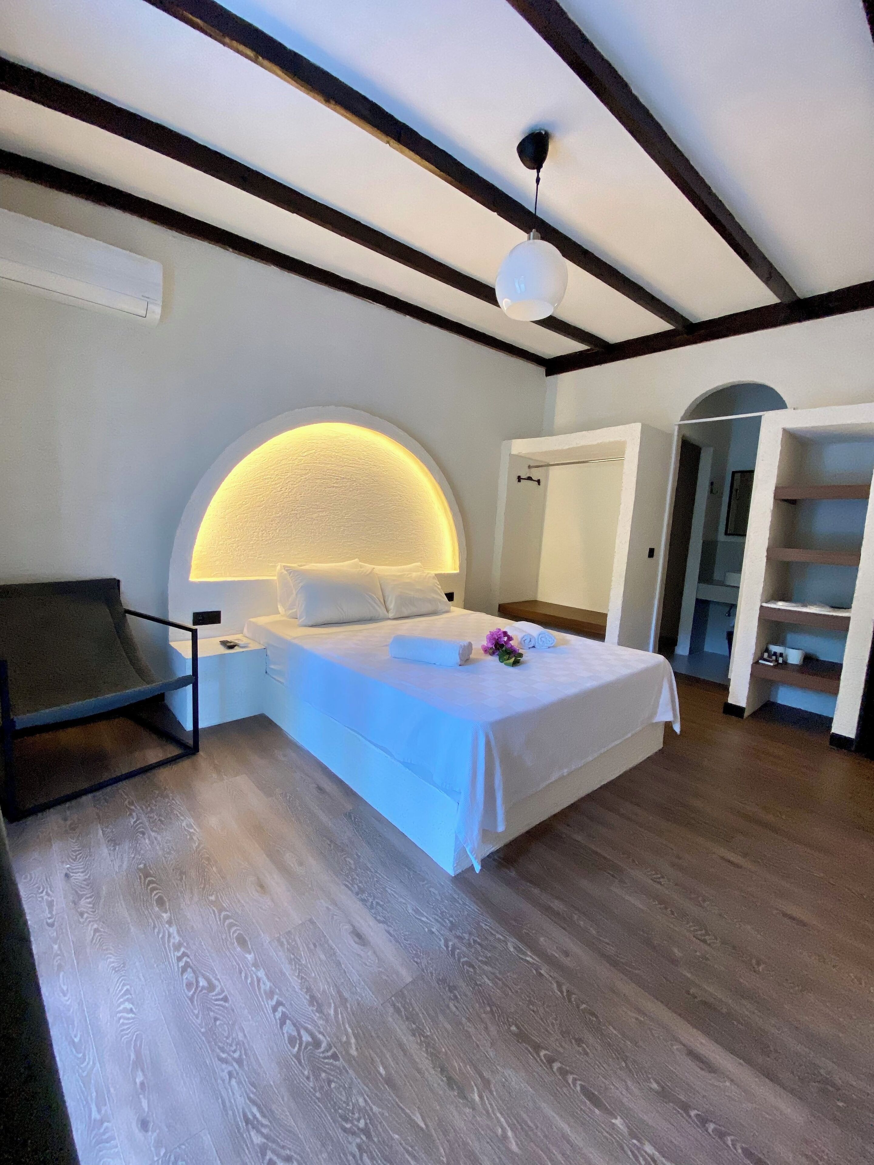 A bedroom with a large bed, a headboard with a built-in light, a chair, and a wooden floor.