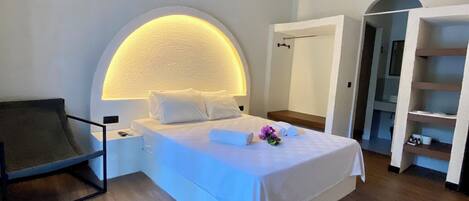 A bedroom with a large bed, a headboard with a built-in light, a chair, and a wooden floor.