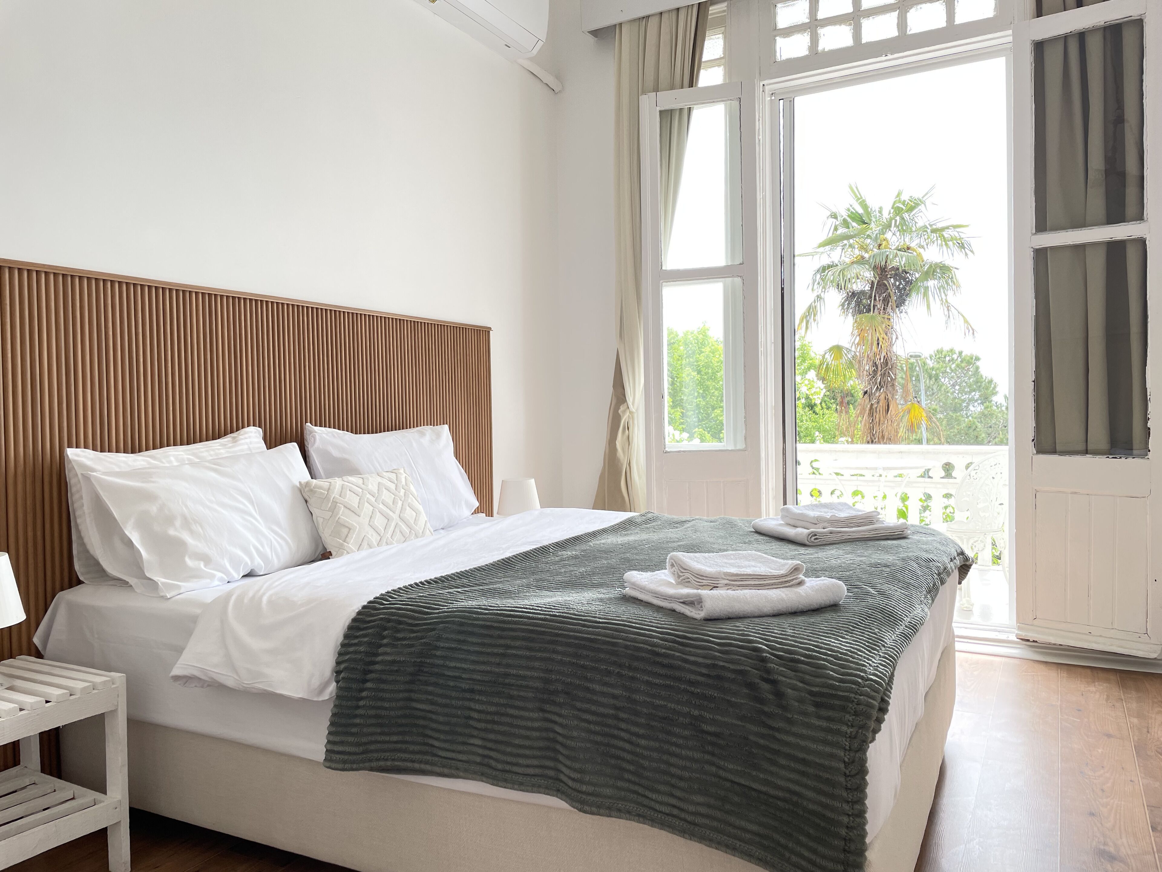 A neatly made bed with white linens and a green striped blanket, a wooden headboard, and a view of greenery through an open window.