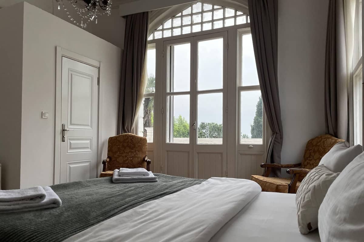 A hotel room with a large bed, two chairs, a window with curtains, and a chandelier.