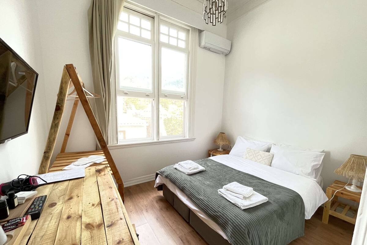 A bedroom with a wooden desk, a bed with a green blanket, a TV mounted on the wall, and a window with curtains.