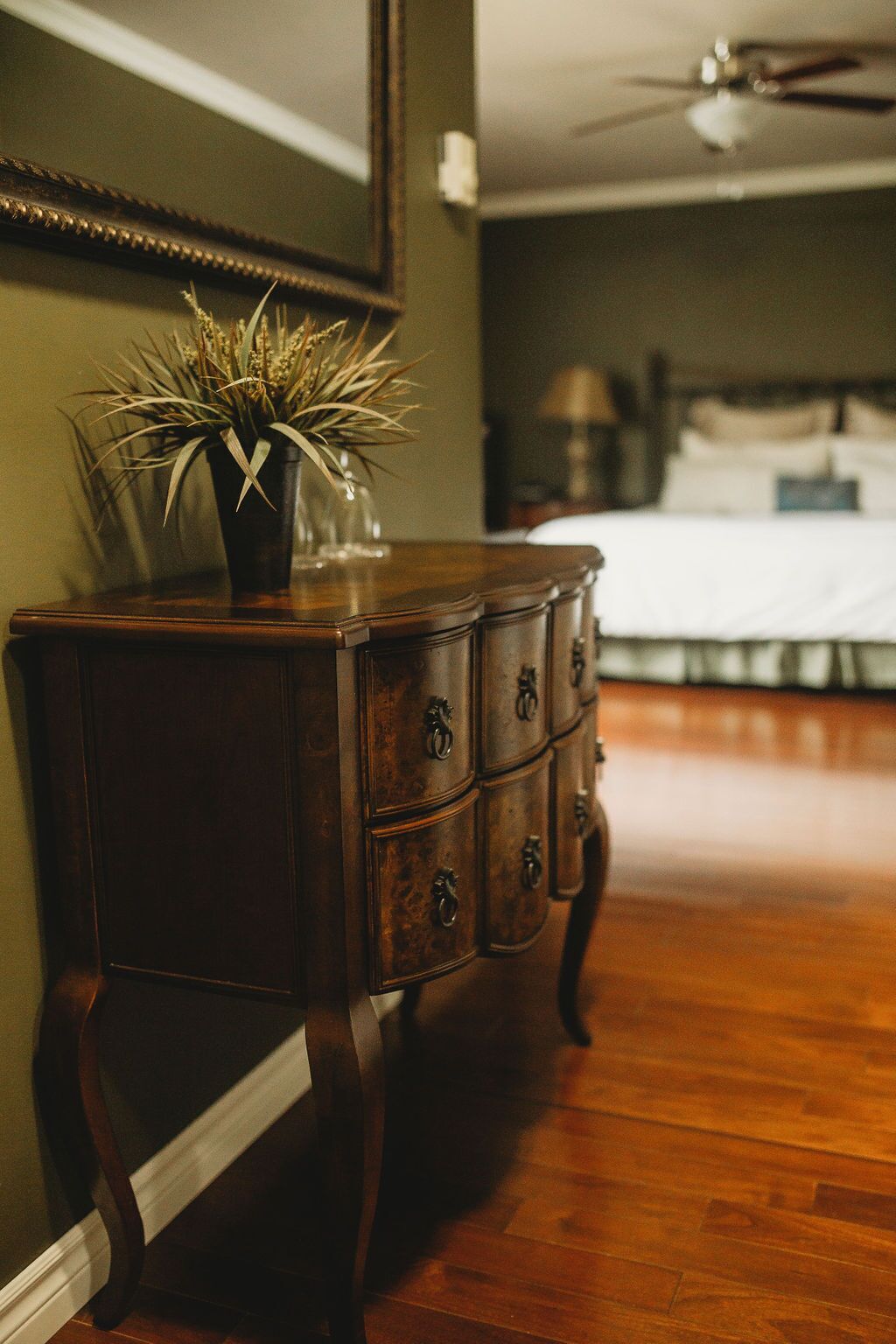 A wooden dresser with drawers, a vase with dried plants, and a mirror on the wall.