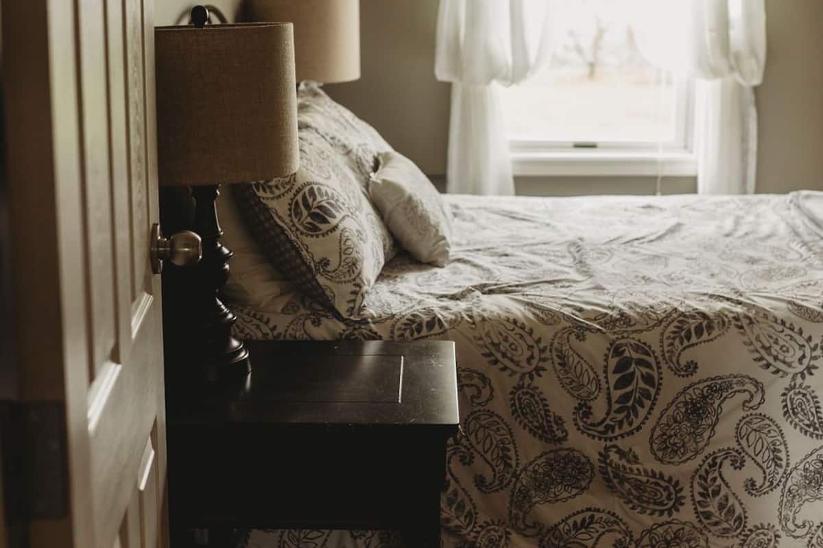 A bedroom with a bed, a bedside table with a lamp, a window with curtains, and a door.