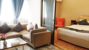 A modern hotel room with a sofa, a small coffee table with a vase of flowers, a bed with a headboard, and a sliding door leading to another room.