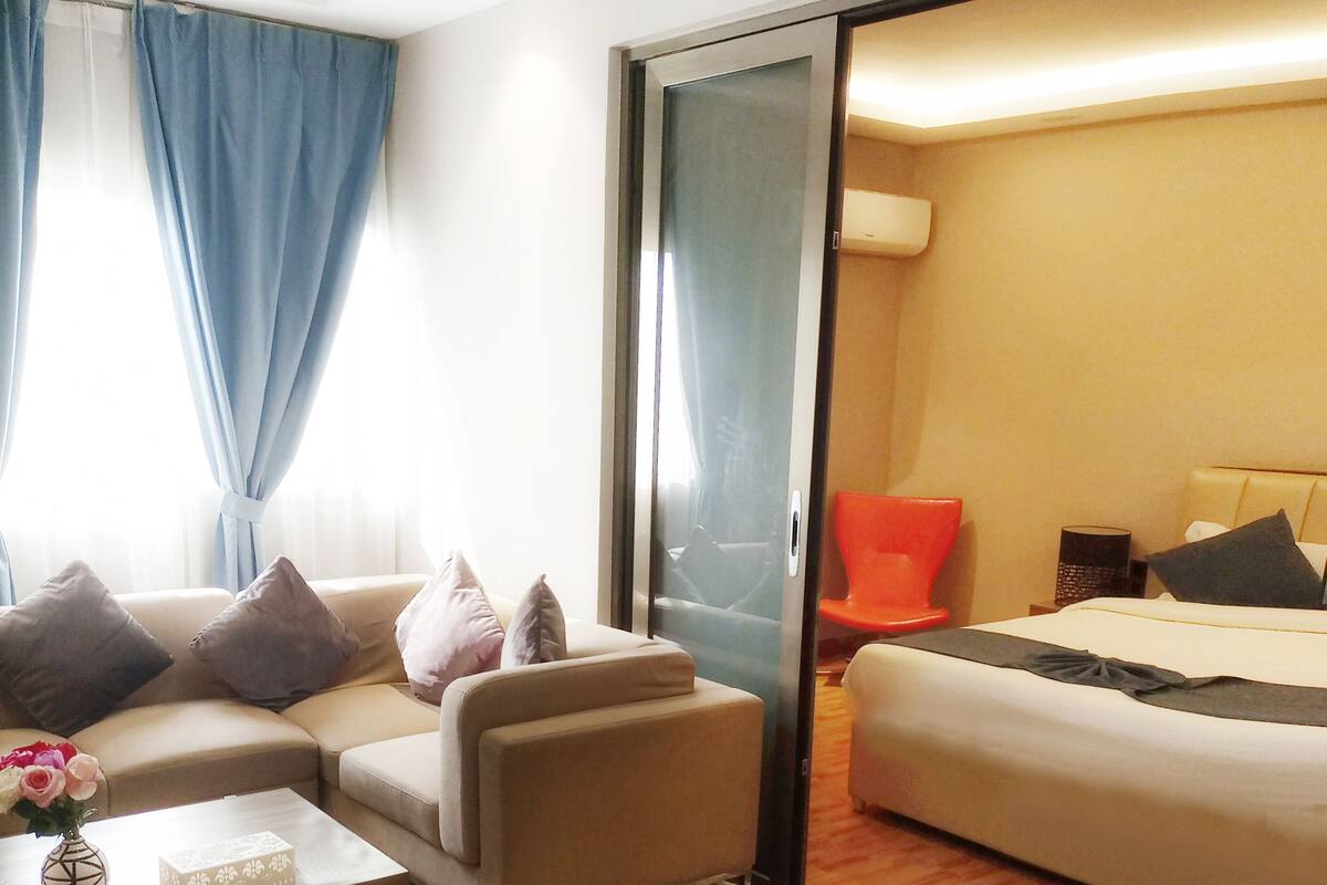 A modern hotel room with a sofa, a small coffee table with a vase of flowers, a bed with a headboard, and a sliding door leading to another room.