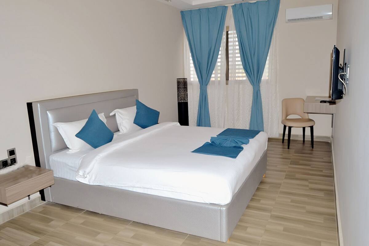 A neatly made bed with white linens and blue pillows, a bedside table with a lamp, a chair, and a desk with a computer.