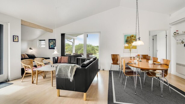 A modern living room with a black sofa, a dining table with chairs, and a large window providing a view of greenery. - Amazing home in Brovst with kitchen (Brovst)