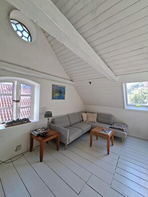 Una habitación tipo loft acogedora con techo inclinado, un sofá gris, dos mesitas de madera y una ventana con vista a un tejado de tejas rojas.