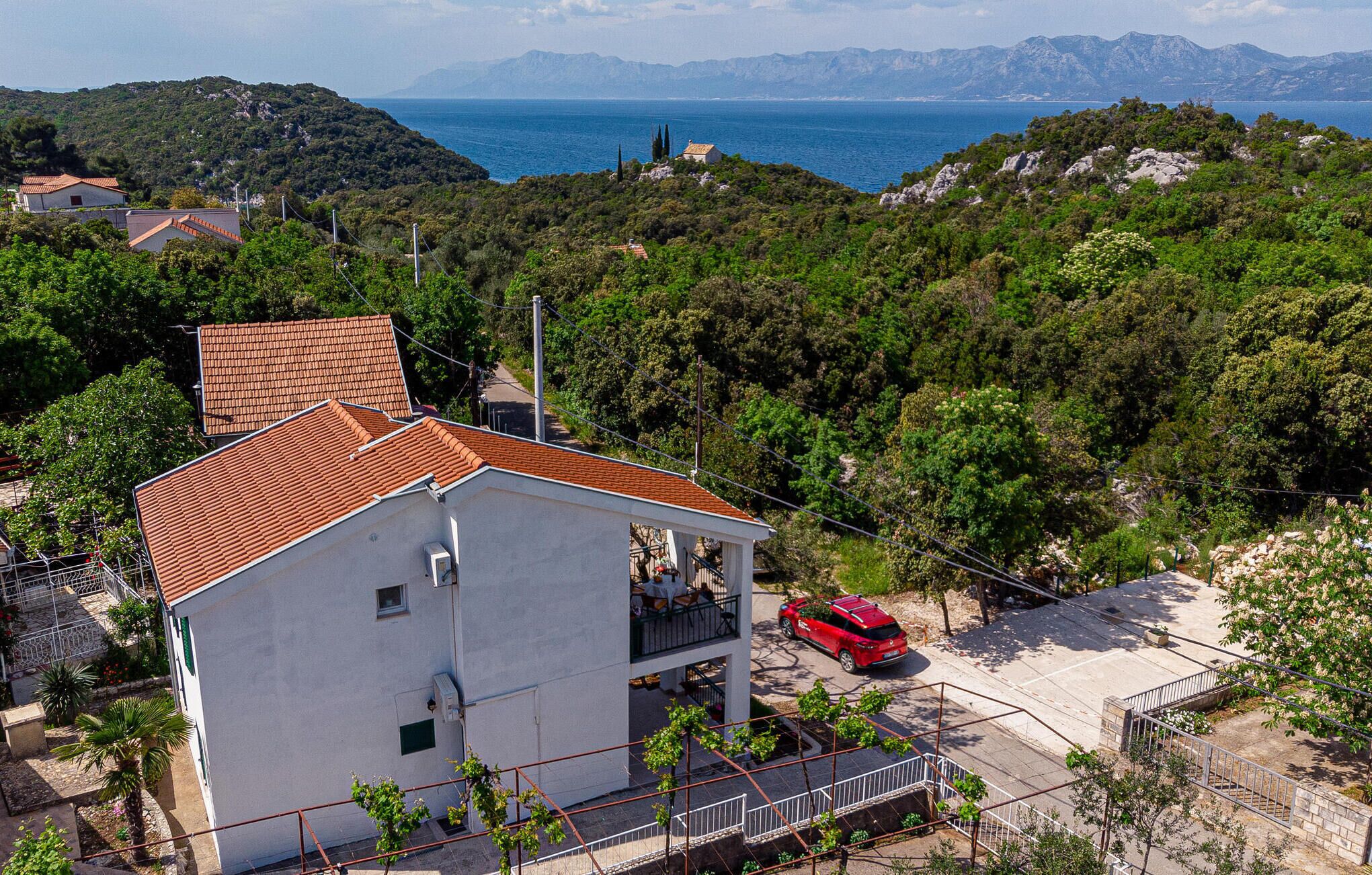 Un bâtiment blanc avec un toit recouvert de tuiles rouges, une voiture rouge garée devant, et une vue sur la mer et les montagnes en arrière-plan.