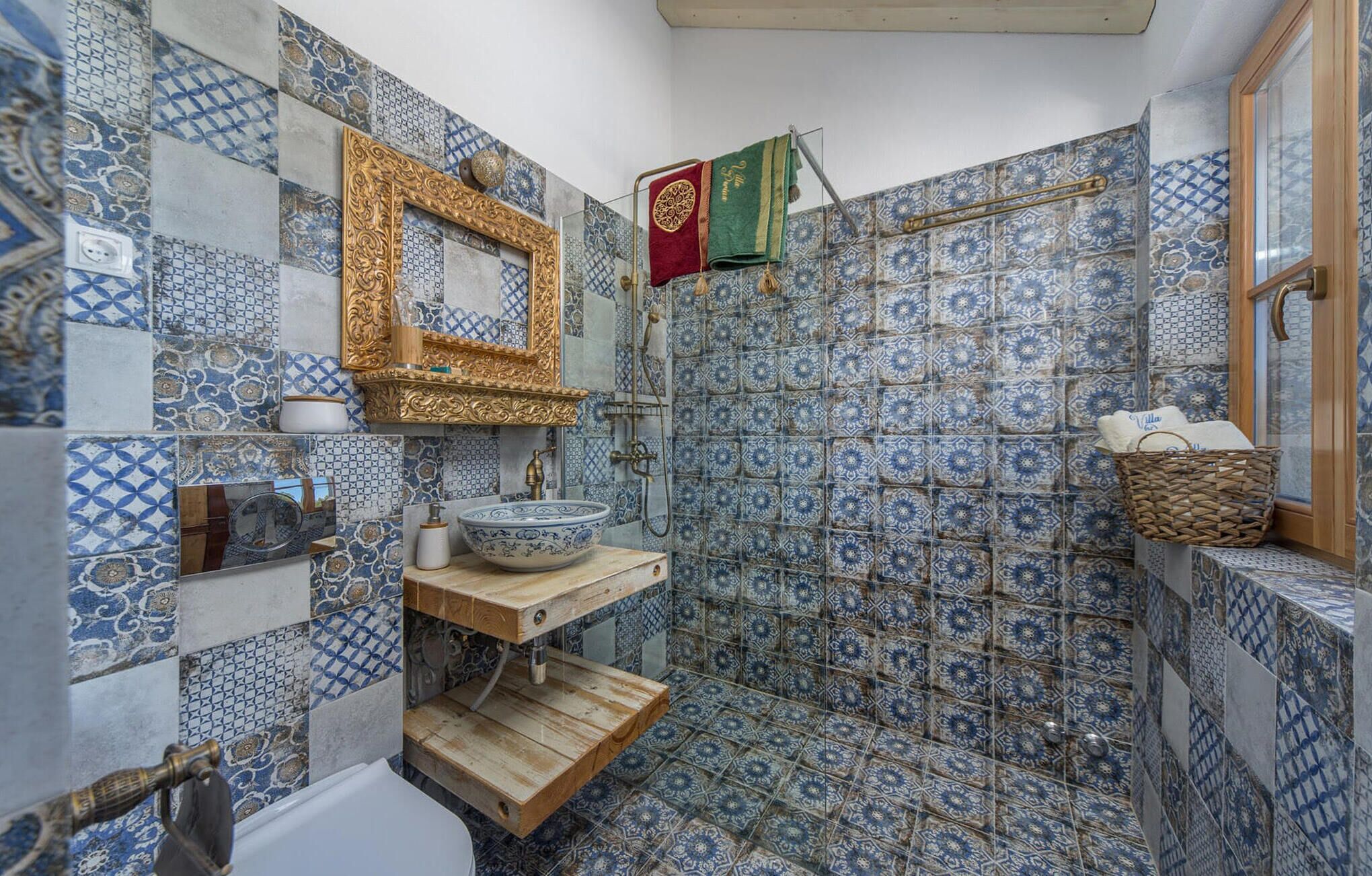 A bathroom with blue and white patterned tiles, a wooden vanity, a gold-framed mirror, and a hanging towel rack.