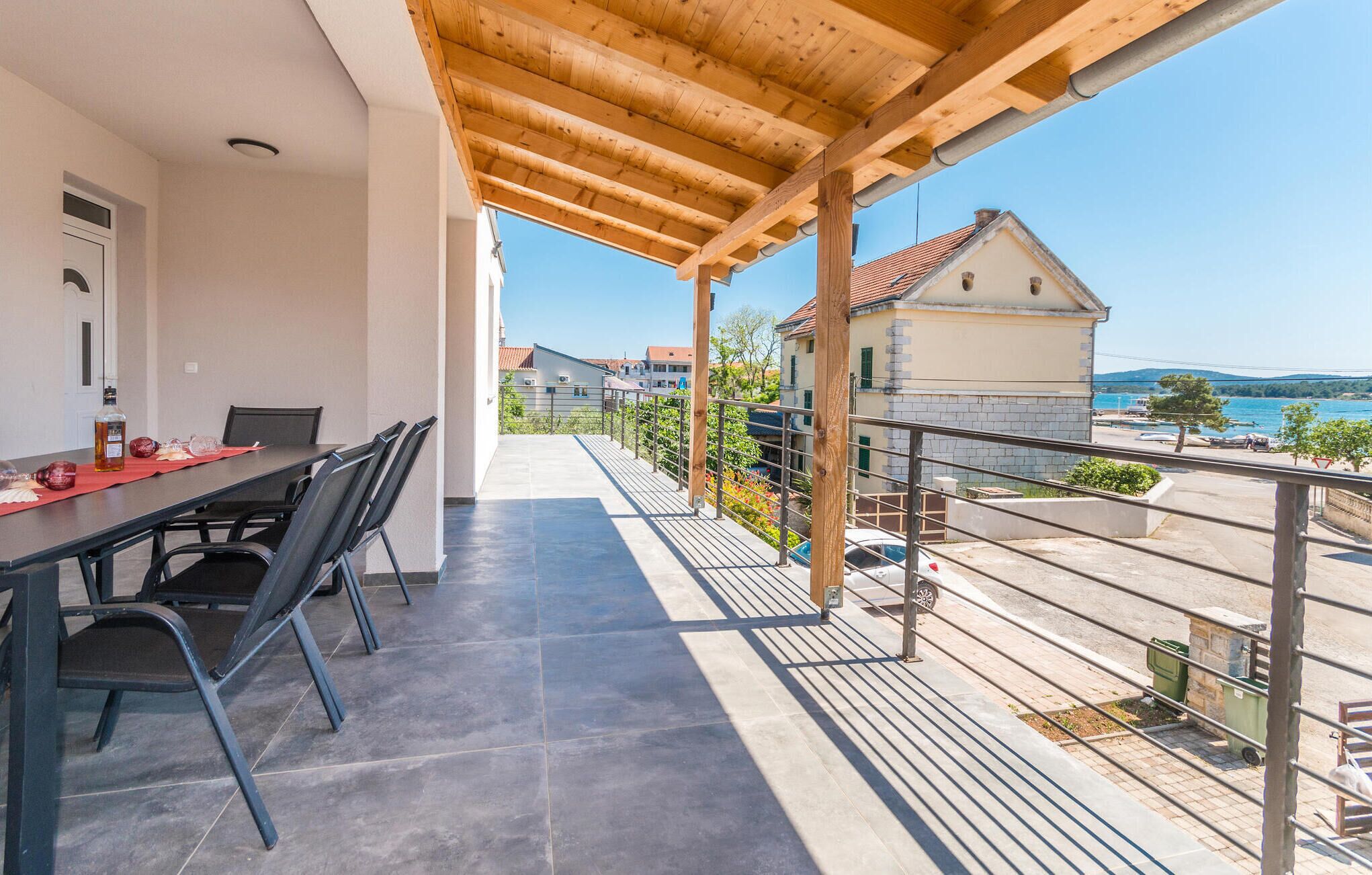 A balcony with a table and chairs, overlooking a coastal area with buildings and a clear sky.
