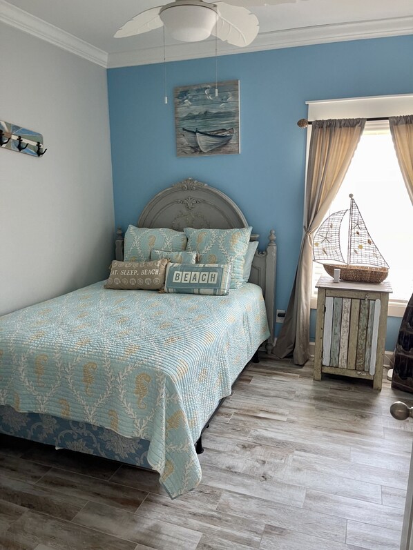 A bedroom with a bed, a ceiling fan, a painting, a window with curtains, and a small table with a decorative item.