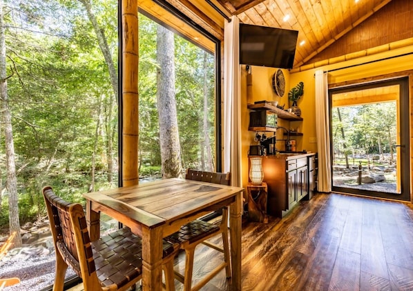 Ein Holzinterieur mit Esstisch und Stühlen, einer Küchenzeile mit Spüle und Schränken sowie großen Fenstern mit Blick auf den Wald.
