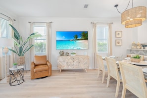 Ein Wohnzimmer mit einem großen Fernseher, der eine Strandansicht zeigt, einem braunen Ledersessel, einem Holzschrank mit Fischgrätmuster und einem Essbereich mit Holztisch und -stühlen.