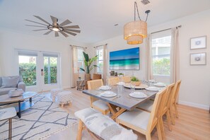 Un coin repas avec une table dressée pour quatre personnes, des chaises en bois, un ventilateur de plafond et une grande fenêtre avec des rideaux.
