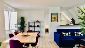 Ein modernes Wohnzimmer mit einem blauen Sofa, einem Esstisch mit violetten Stühlen und einer Küchenzeile mit Regalen und einem roten Topf.