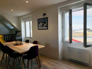 Een eethoek met een houten tafel en stoelen, een telescoop op een statief, en een groot raam met uitzicht op het strand en gebouwen.
