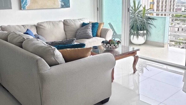 A modern living room with a beige sofa, a coffee table, and a large window offering a view of a cityscape.