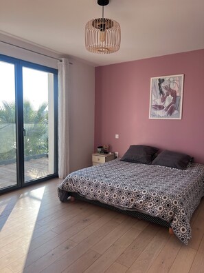 A bedroom with a large bed, a sliding glass door to a balcony, a ceiling light, and a painting on the wall.